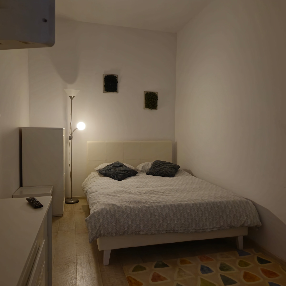 A softly lit bedroom features a spacious bed with patterned bedding accented by two pillows. A white lamp casts gentle light, enhancing the room's calm atmosphere. Decorative wall art is visible above the bed, contributing to the minimalist design.