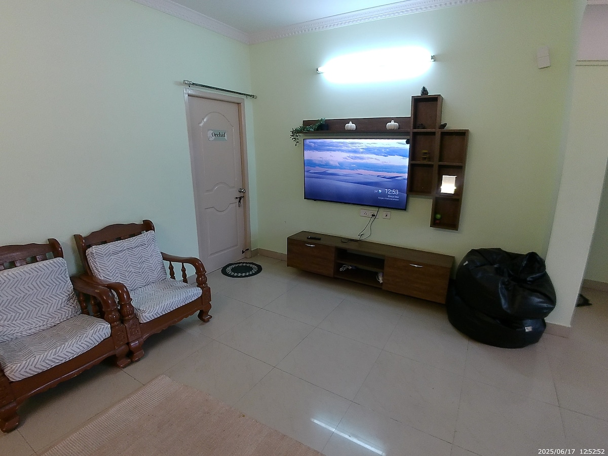 Cozy living room with a flat-screen TV, comfy wooden chairs, and a bean bag. Bright, clean, and perfect for relaxing with family after a long day.