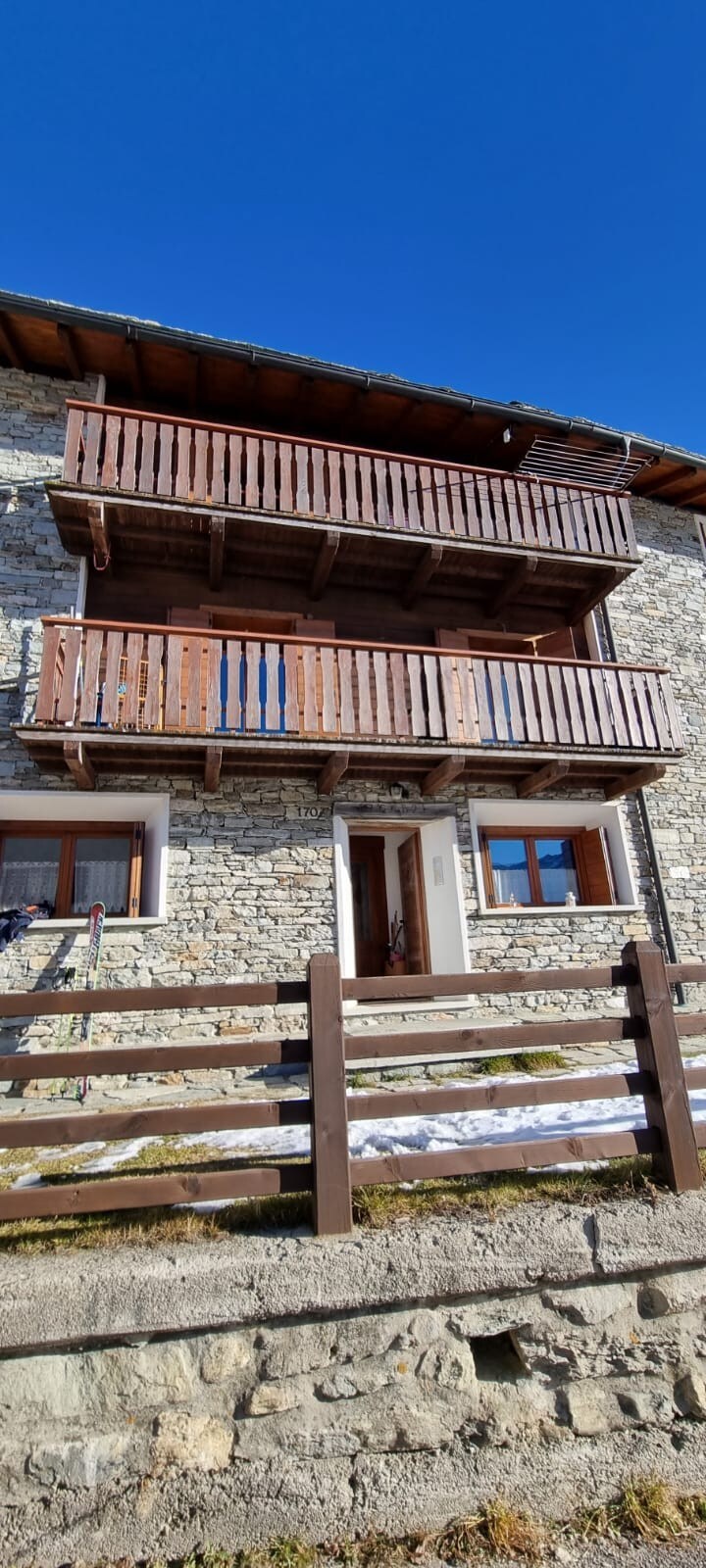The exterior of the stone building features two wooden balconies providing outdoor space. The entrance is framed by two windows on either side, and a wooden fence borders the area, adding to the structure's rustic charm.