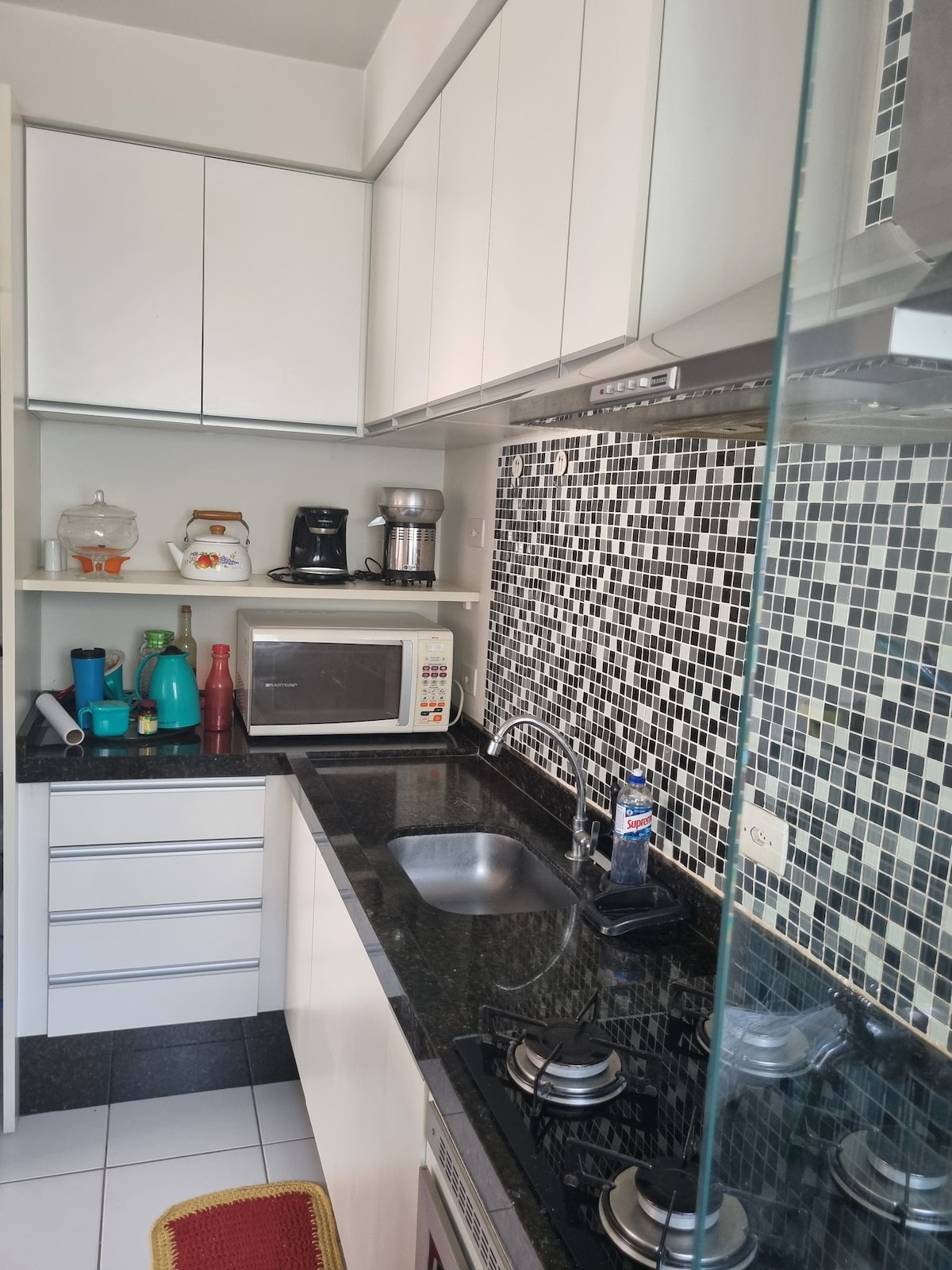 The kitchen is equipped with modern appliances, including a microwave, coffee maker, and a cooktop with several burners. Black granite countertops complement the mosaic tile backsplash, while white cabinetry offers storage space. A sink is positioned conveniently near the cooking area.