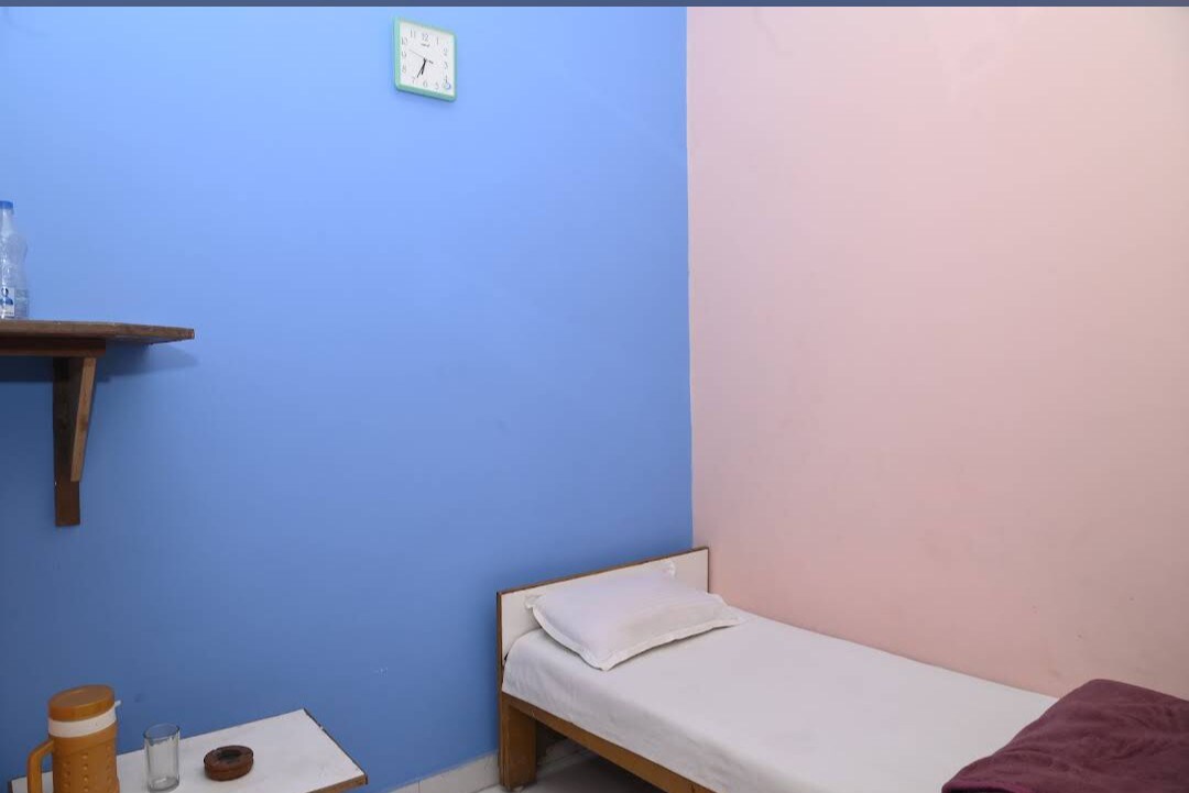 A simple room features a single bed with a white mattress and a pillow. The walls are painted in pastel blue and pink. A small shelf holds a glass and a wooden bowl, while a clock is mounted above the bed.
