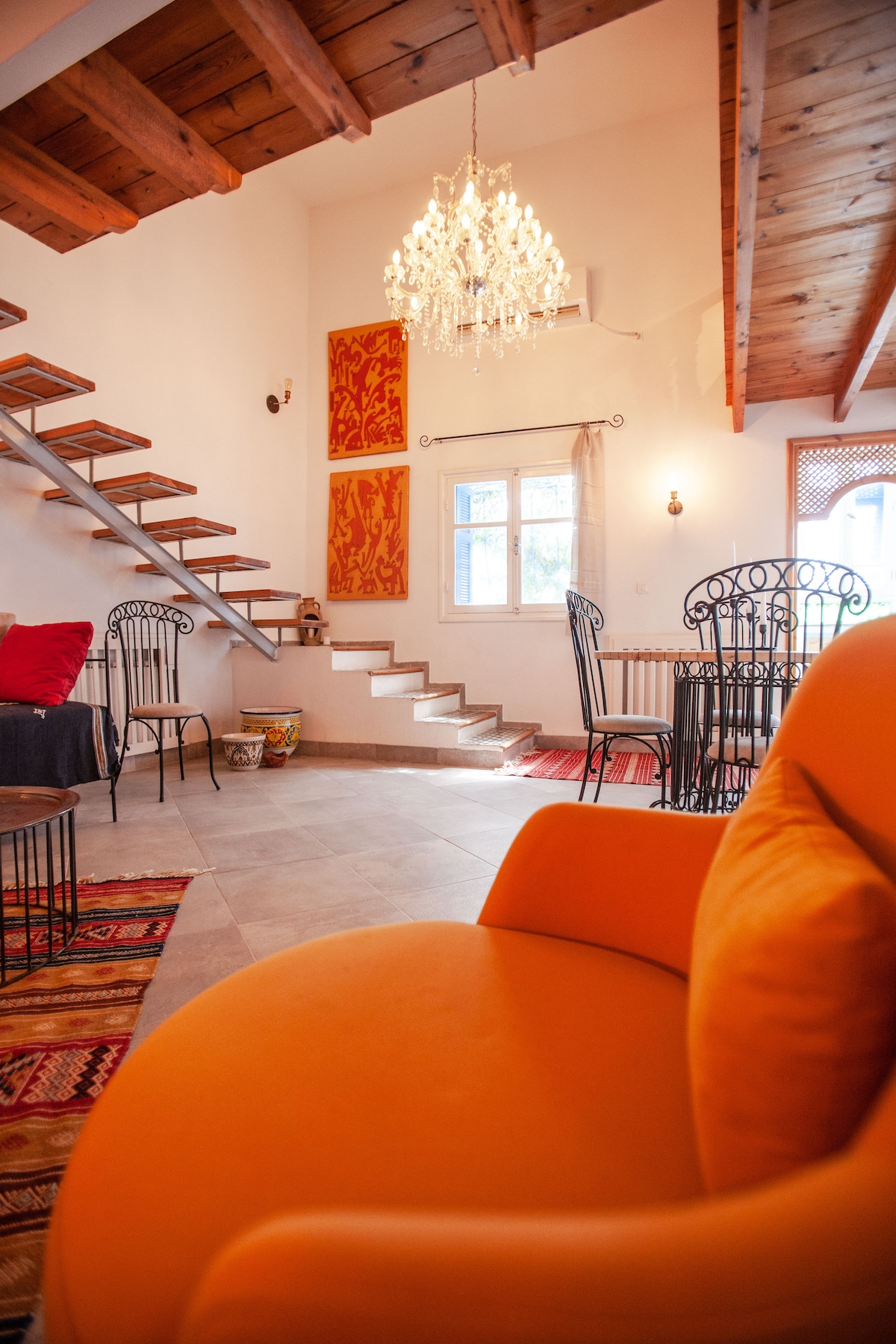 An inviting living area features a bright orange chair in the foreground, with traditional artwork displayed on the walls. A staircase leads to the upper floor, and a chandelier hangs from the ceiling. A dining table with chairs is positioned near a window, allowing natural light.