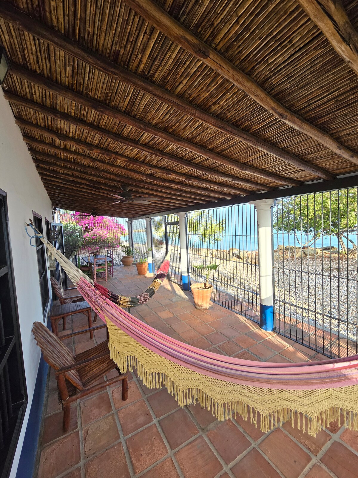 A covered terrace features two colorful hammocks draped between posts, providing a relaxing outdoor space. Potted plants and blooming flowers add greenery, while the open view towards the water allows for ocean breezes. Textured tiles create a warm, inviting ground surface.