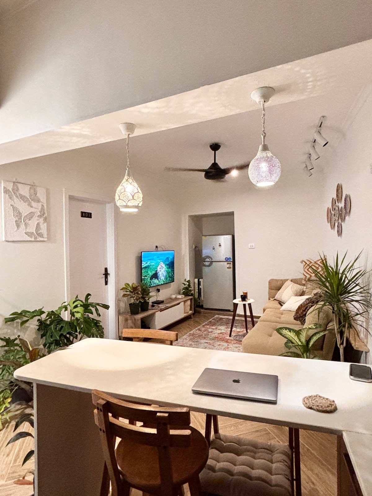 A cozy living area is highlighted by light-colored walls and a comfortable sofa, accompanied by decorative plants. Soft pendant lights dangle overhead, illuminating a small dining table. A TV is positioned against the wall, creating a relaxed space for gatherings or quiet moments.