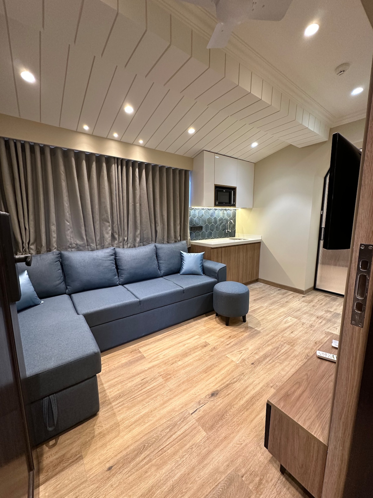 A cozy living room area features a modern sectional sofa in a soft blue hue, complemented by a matching ottoman. Warm wood flooring enhances the inviting atmosphere. In the background, a sleek kitchenette is partially visible, equipped with stylish cabinetry and appliances.