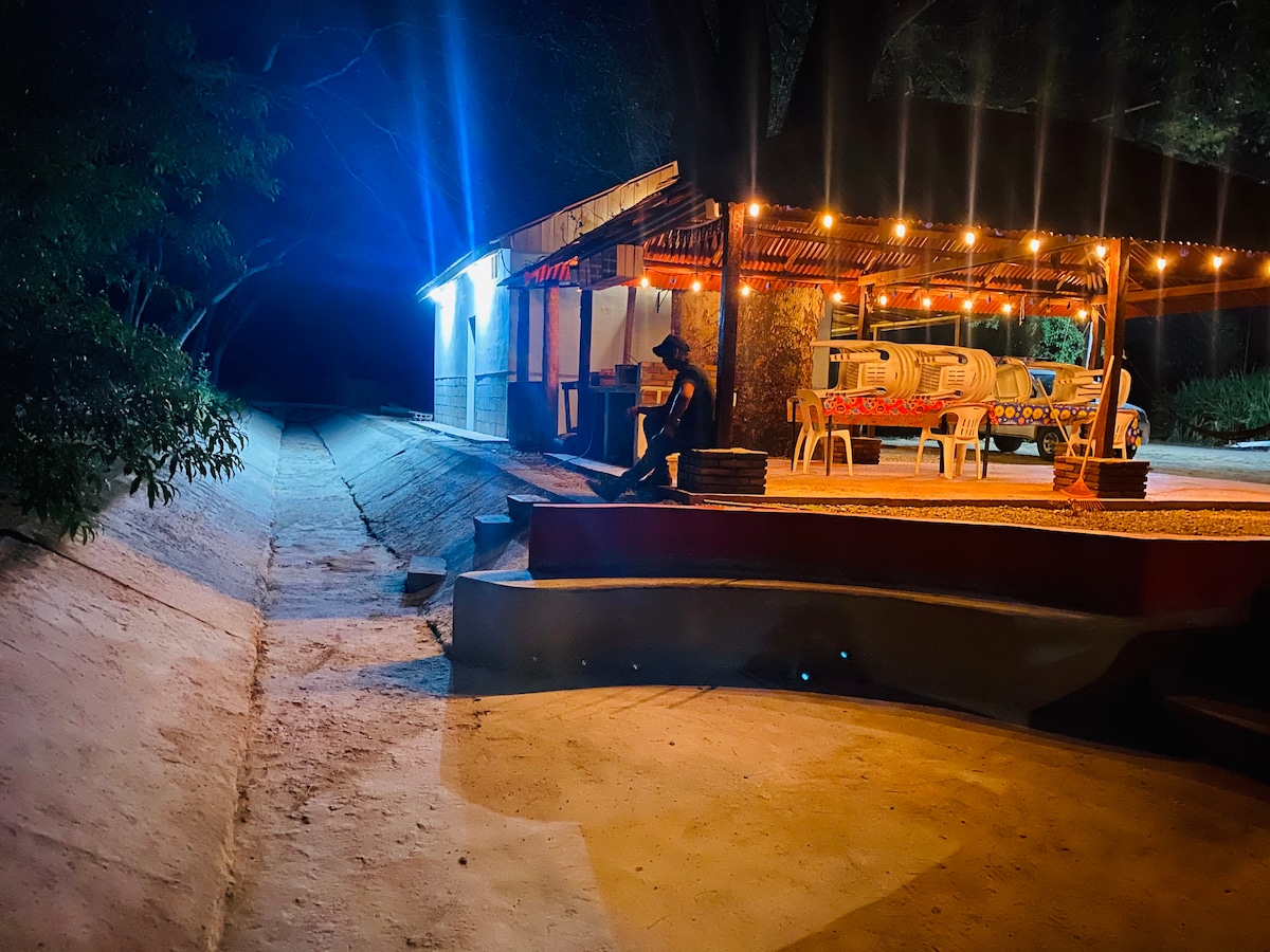 An outdoor seating area is illuminated by string lights, creating a cozy environment in the evening. A path leads away from the structure, flanked by greenery and a softly lit backdrop, inviting calm and relaxation.