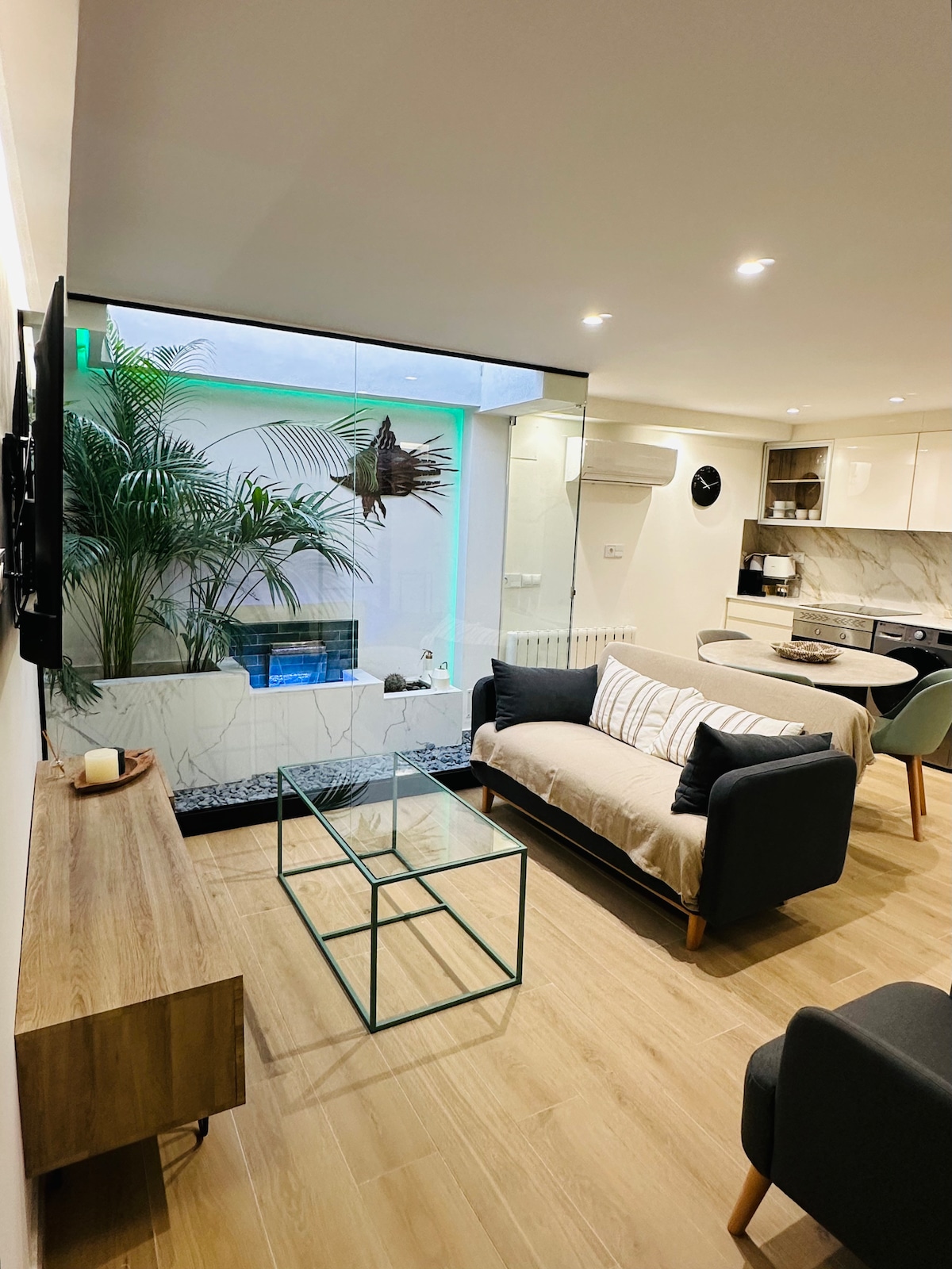 A modern living area features a comfortable sofa and accent chairs positioned around a glass coffee table. Natural light fills the space, highlighting a stylish kitchen in the background. An interior patio with greenery and a small water feature adds a serene touch to the atmosphere.