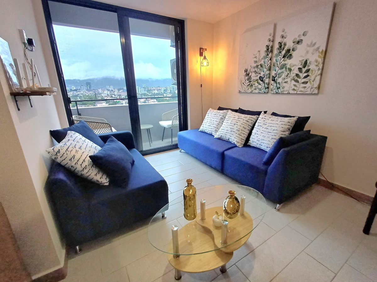 A modern living area features a large blue sofa and a smaller blue seating option. A glass table sits in front, adorned with decorative items. Floor-to-ceiling windows provide a view of the cityscape, while light-colored walls are complemented by botanical artwork.