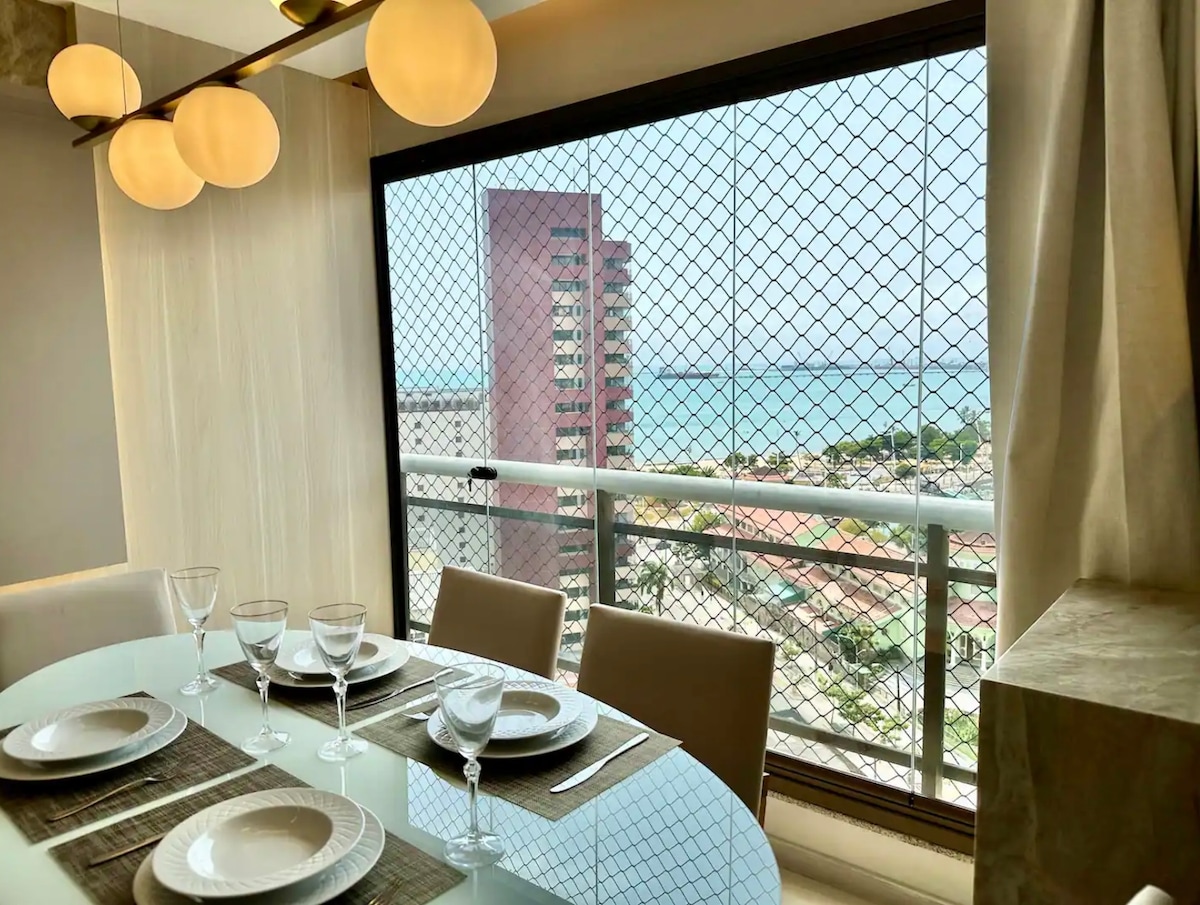 A dining area is visible, featuring a round glass table set with plates and elegant glassware. Large windows showcase a scenic view overlooking the ocean and city, with a protective mesh screen. Soft light emanates from distinctive hanging lamps above the table.