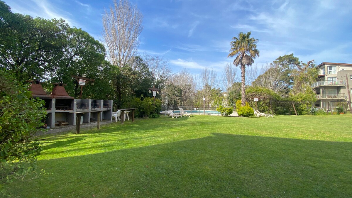 A large green park is depicted, featuring a swimming pool in the background surrounded by lush trees. Two barbecue grills are present, alongside several lounge chairs scattered on the well-maintained lawn, creating an inviting outdoor space for relaxation and leisure.