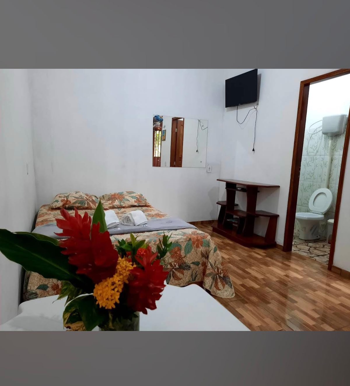 A bright sleeping area features a floral-patterned bedspread over a spacious bed. A wooden entertainment unit is positioned against the wall, while a small table with a vase of colorful flowers adds a touch of decor. The adjoining bathroom is visible through an open door.