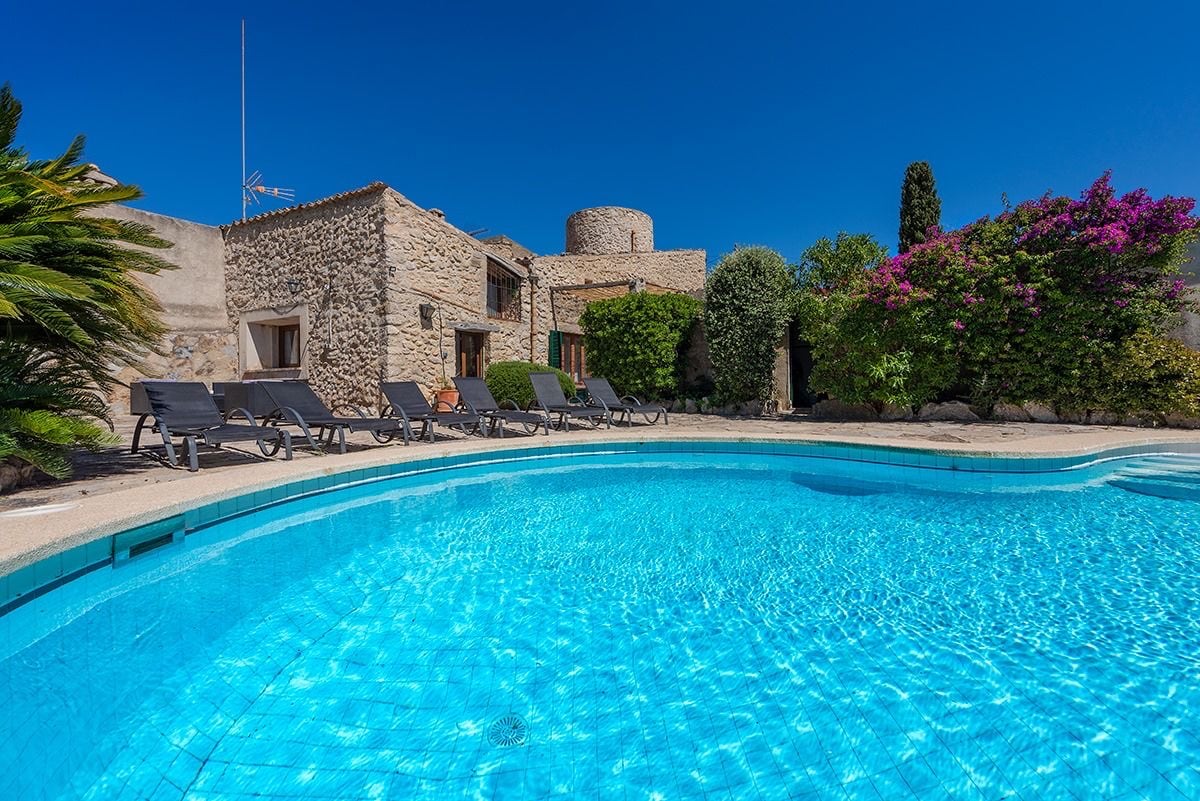 A large swimming pool is visible, featuring clear turquoise water and surrounded by a sun terrace with multiple lounge chairs. The villa's stone facade is presented in the background, with lush greenery and blooming flowers enhancing the serene outdoor space.