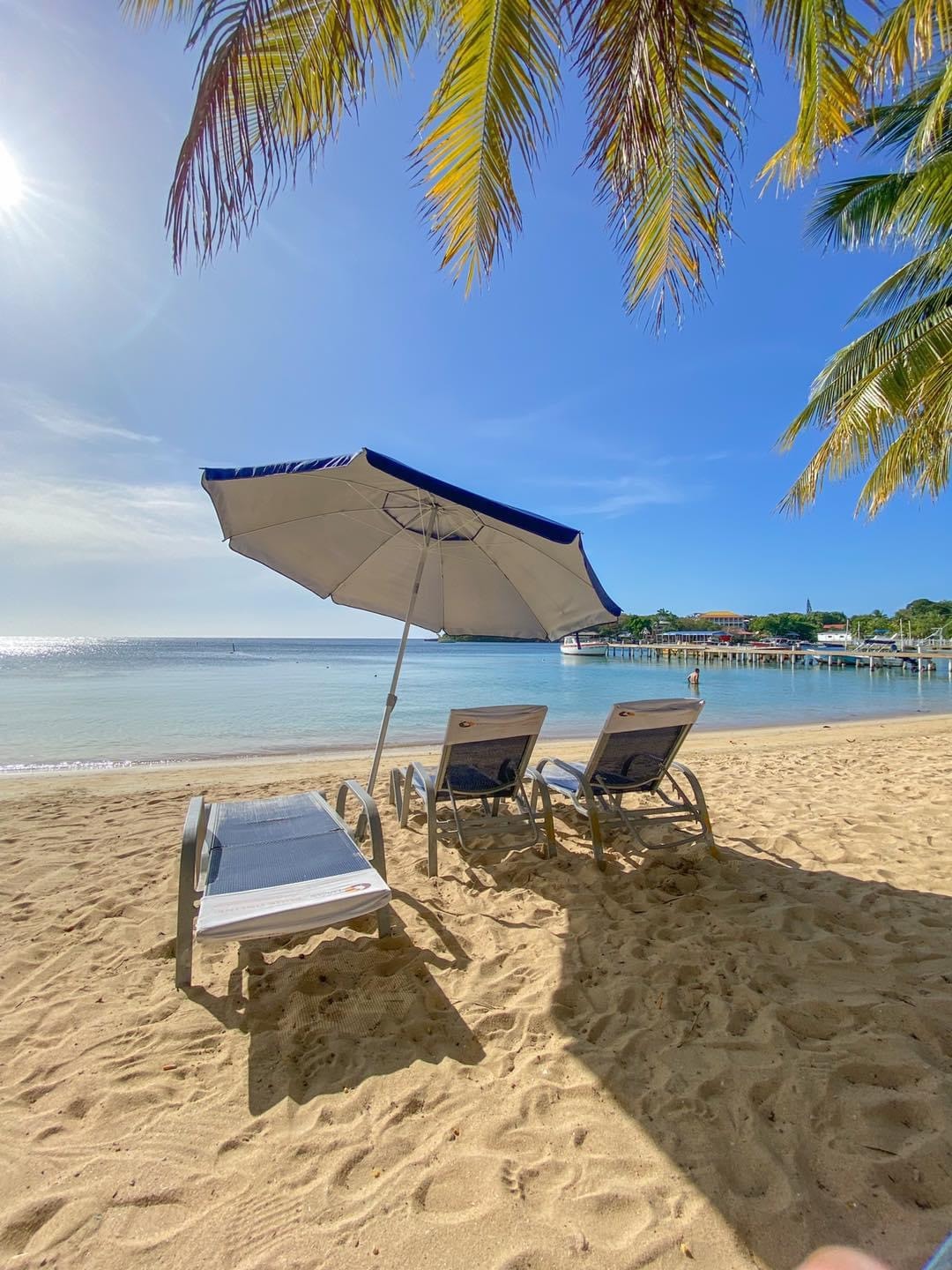 Two lounge chairs sit on soft sandy beach under a large umbrella, positioned to overlook calm waters. The sun shines brightly in a clear blue sky, creating a serene atmosphere. Palm leaves frame the scene, adding a touch of tropical ambiance.