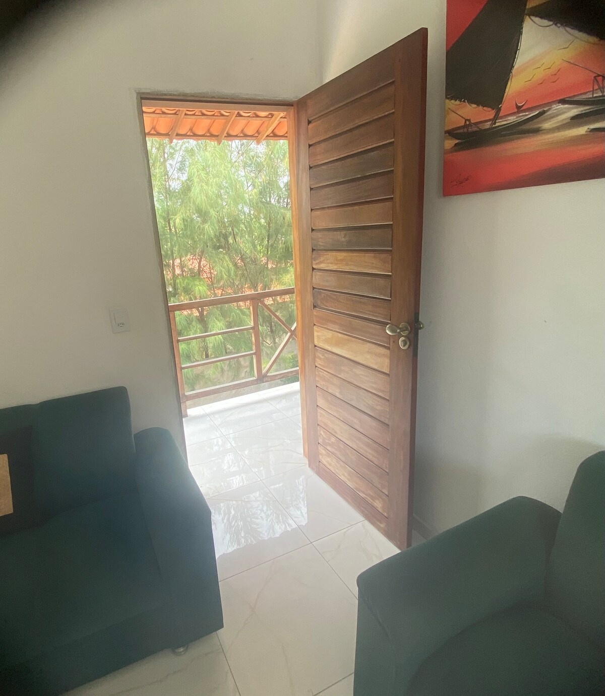 A wooden door is positioned slightly ajar, revealing a bright outdoor balcony with a view of greenery. Two green sofas are visible within the inviting room, complemented by a colorful wall art piece featuring a nautical scene.