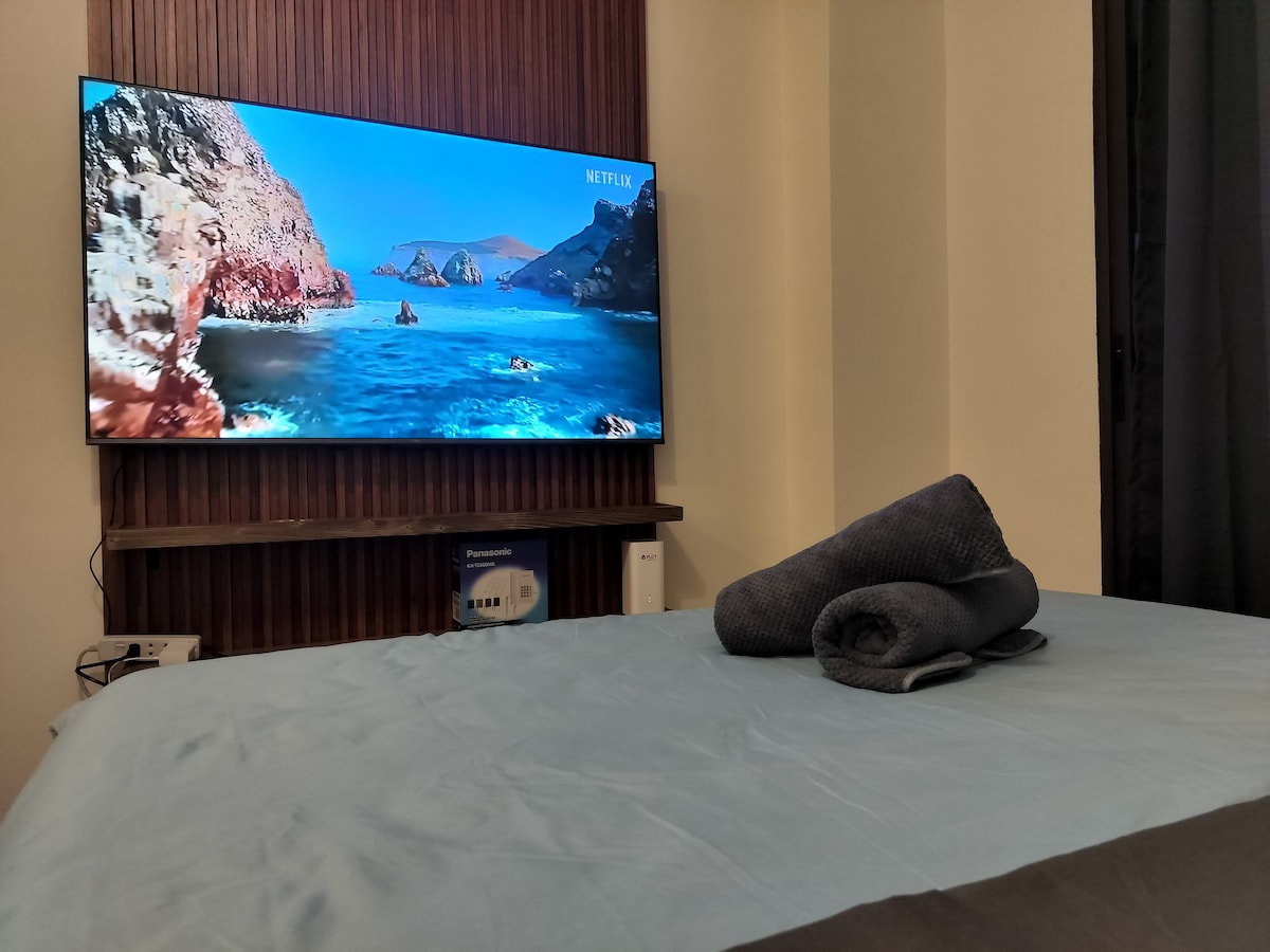 A spacious bed is outfitted with light blue linens, accompanied by a neatly rolled dark towel. A large flat-screen television is mounted on the wall, displaying a scenic coastal view. A dark curtain partially covers a window, contributing to the calming ambiance.