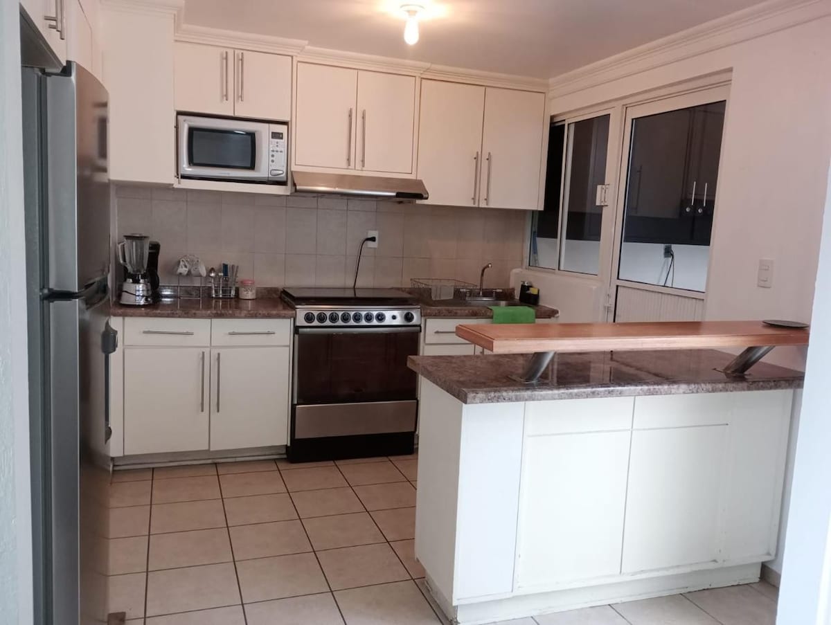 A modern kitchen featuring white cabinetry and granite countertops is equipped with stainless steel appliances, including an oven and microwave. A central island with a wooden countertop offers additional workspace, and large windows allow natural light to brighten the space.