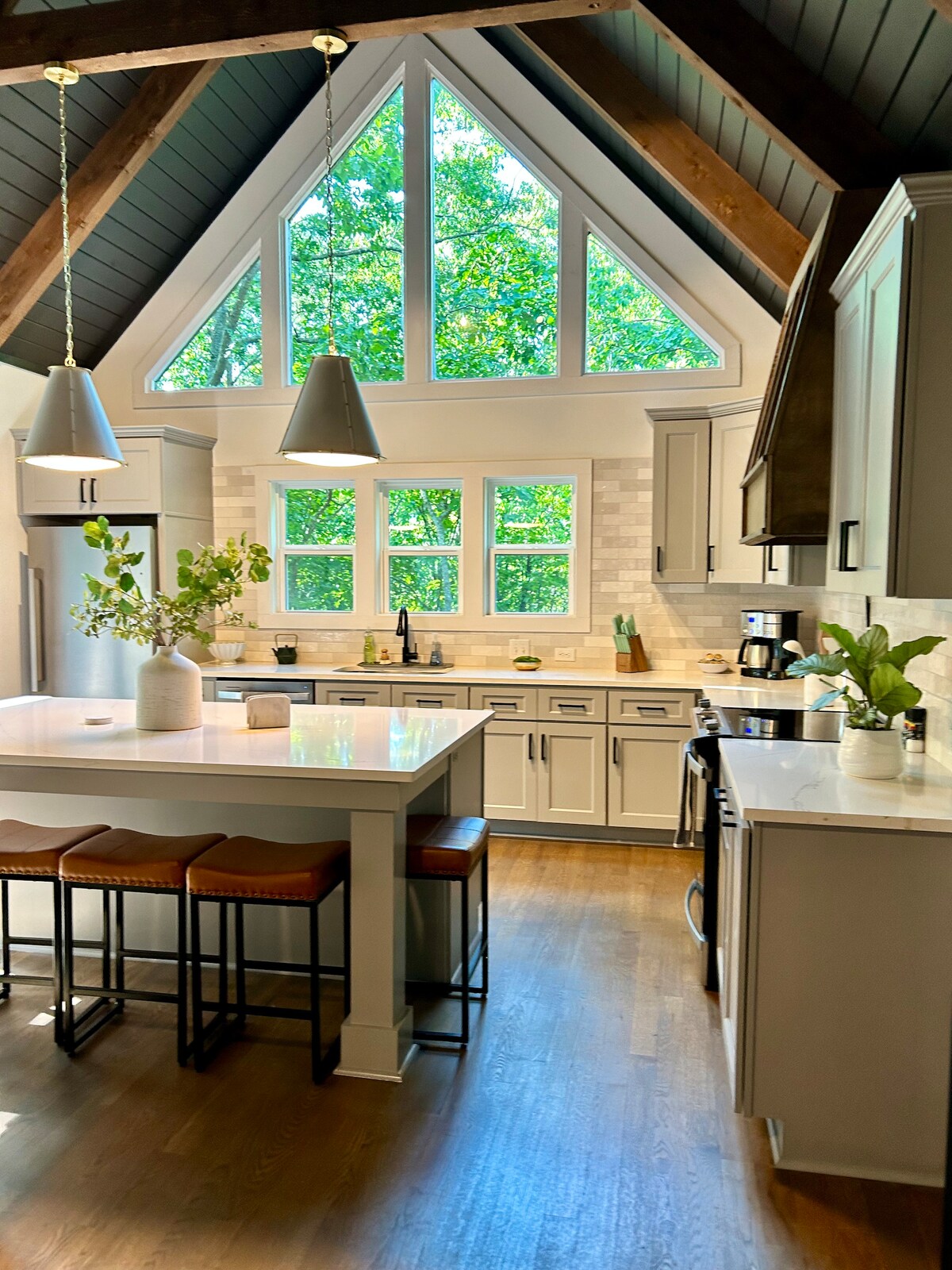A modern kitchen features a spacious island with seating for three. Large windows allow natural light to fill the room, showcasing the wood beams above. Light-colored cabinetry complements the stylish countertops, while greenery enhances the inviting atmosphere.
