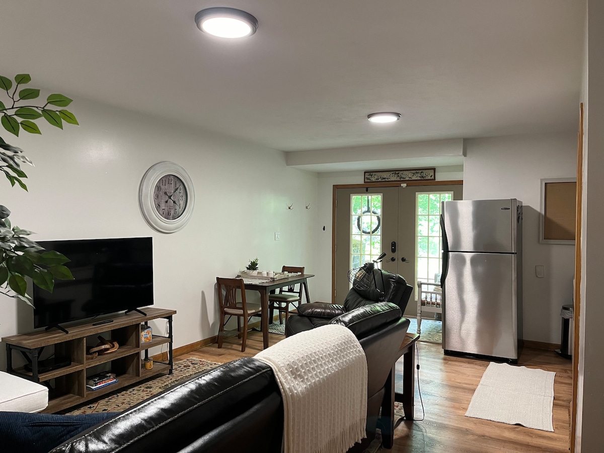 A light and airy living area is visible, featuring a flat-screen television mounted on a wooden stand. A dining table for four sits adjacent to a modern refrigerator. Natural light pours in through the double doors that open to a green outdoor space.