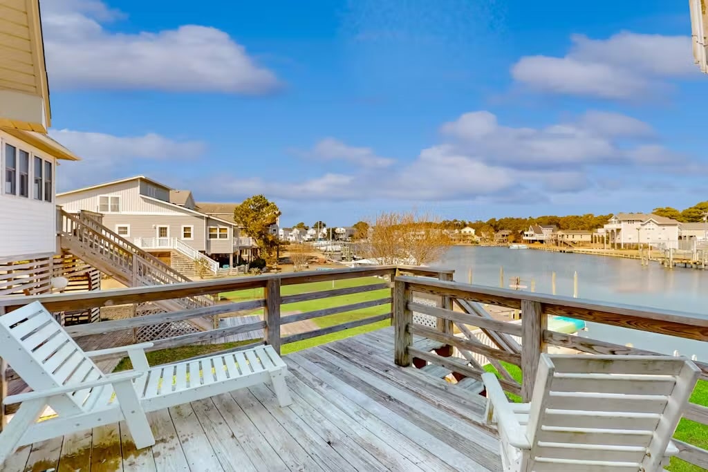 A spacious deck is shown with two white lounge chairs positioned for waterfront views. The calm water is visible, along with neighboring homes in the background. Lush greenery surrounds the area, contributing to the serene outdoor setting.