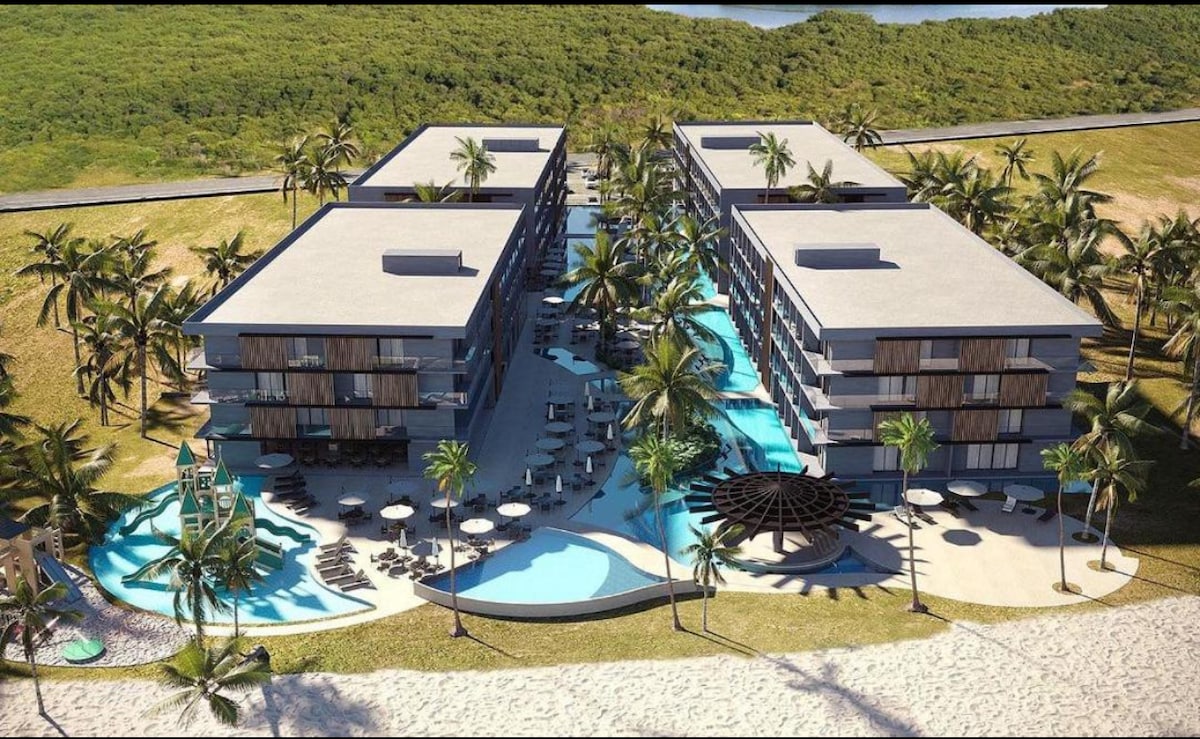 An aerial view reveals a resort complex surrounded by palm trees, featuring interconnected pools and lounging areas. The buildings maintain a modern design with large balconies, while a children's water play area is visible, highlighting a family-friendly atmosphere. The beach is located adjacent to the resort.