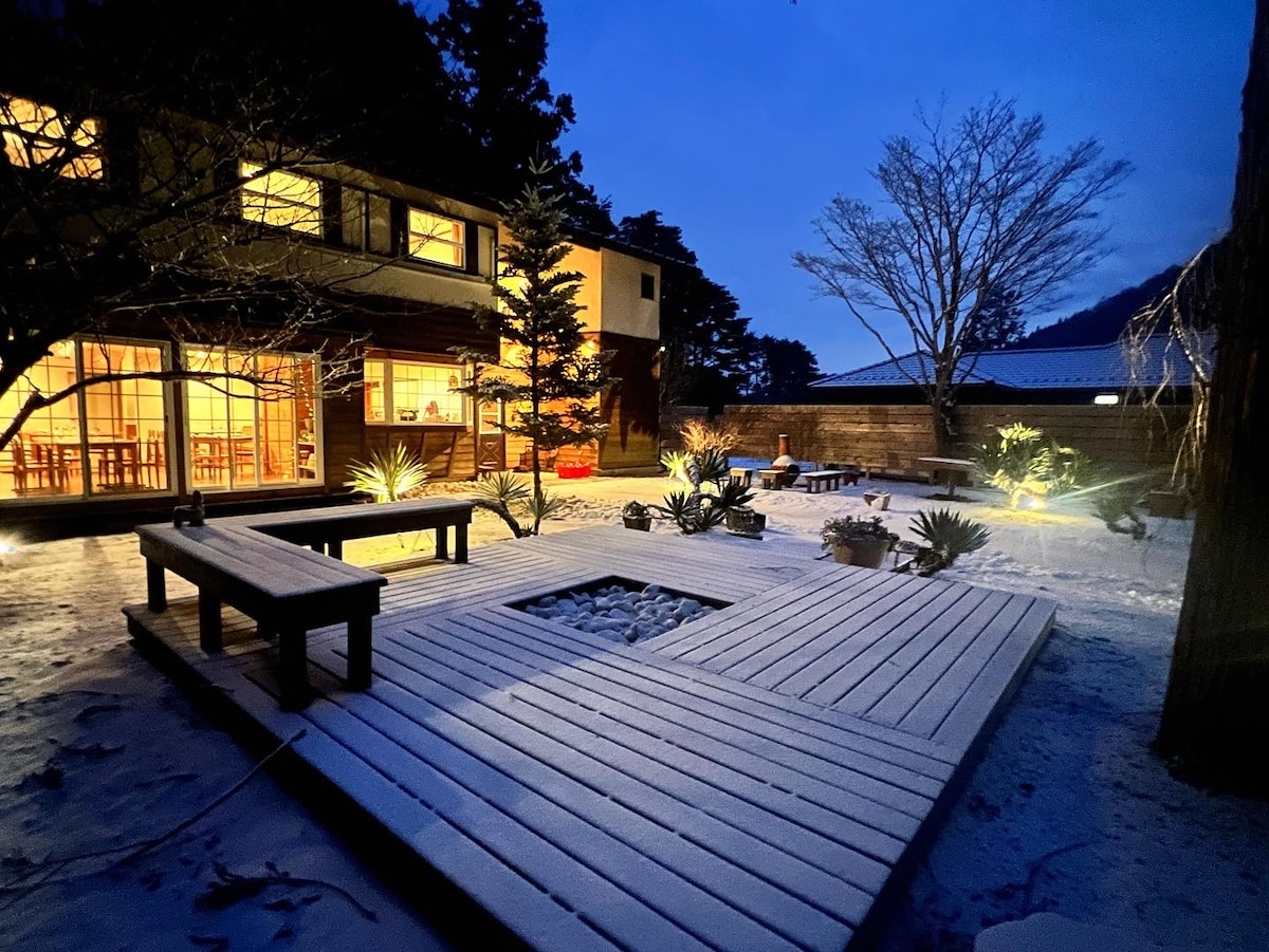 An outdoor deck area is illuminated subtly at dusk, featuring wooden seating and a central square surrounded by gravel. Light glows softly from the windows of the adjacent building, while illuminated plants add a touch of greenery to the winter scene.
