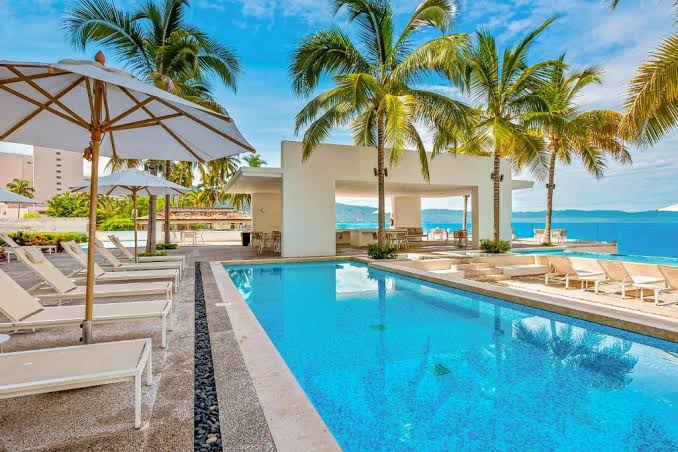 An outdoor pool area is surrounded by palm trees, with comfortable lounge chairs positioned along the edge. A shaded seating area is visible in the background, providing relaxation options. The calm water reflects a clear blue sky, enhancing the serene environment.