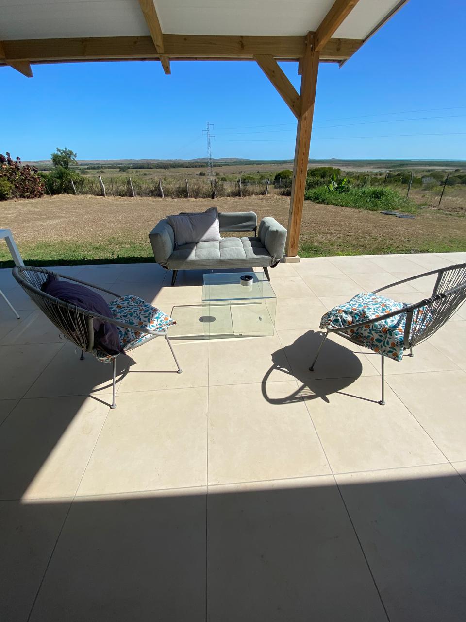An outdoor seating area is featured, with two chairs adorned in colorful fabric and a low glass coffee table. A gray sofa is positioned centrally, offering views of the spacious landscape under a clear blue sky, framed by a wooden structure overhead.