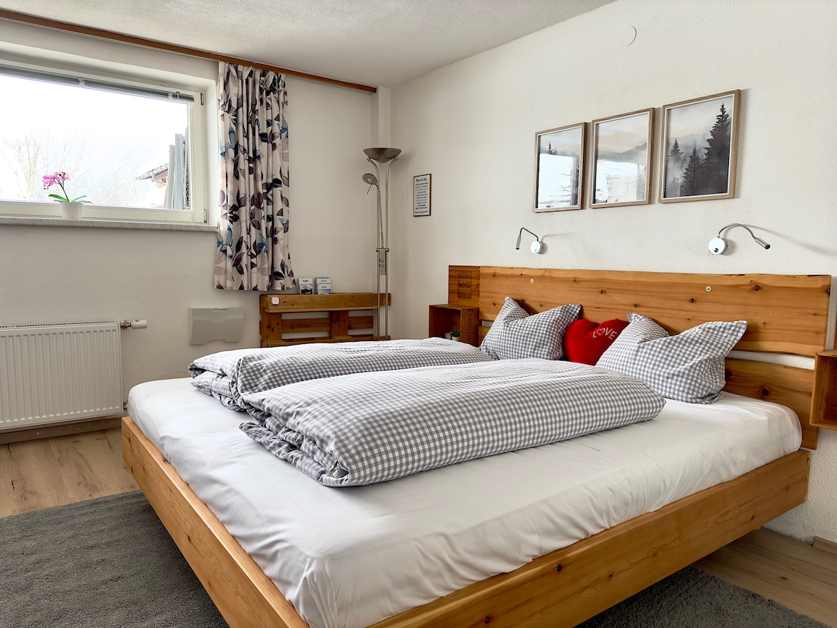 A spacious bedroom features a hand-crafted wooden bed with cozy bedding and inviting pillows. Soft light enters through the window, complemented by patterned curtains. Wall-mounted lamps provide additional lighting, while framed nature-themed art pieces adorn the walls, enhancing the calming ambiance.