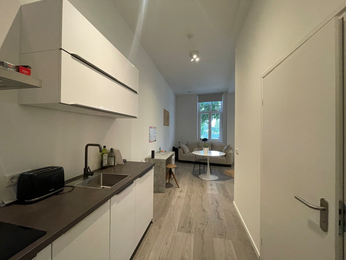 A modern kitchen is situated at the forefront, featuring clean white cabinetry and essential appliances, including a toaster. A dining table is positioned in the center, with a comfortable seating area visible in the background, including a plush sofa and natural light coming from large front windows.