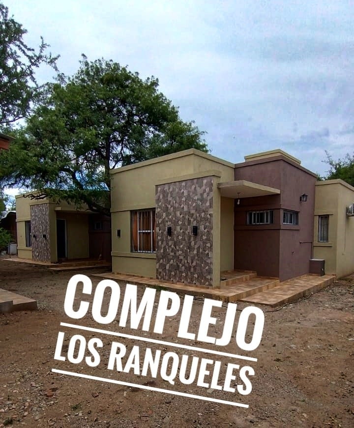 Modern buildings are shown in an outdoor setting, featuring a blend of neutral and earthy tones. Large windows provide natural light, and decorative stone accents contribute texture to the architecture. A gravel path leads to the entrance of the complex.
