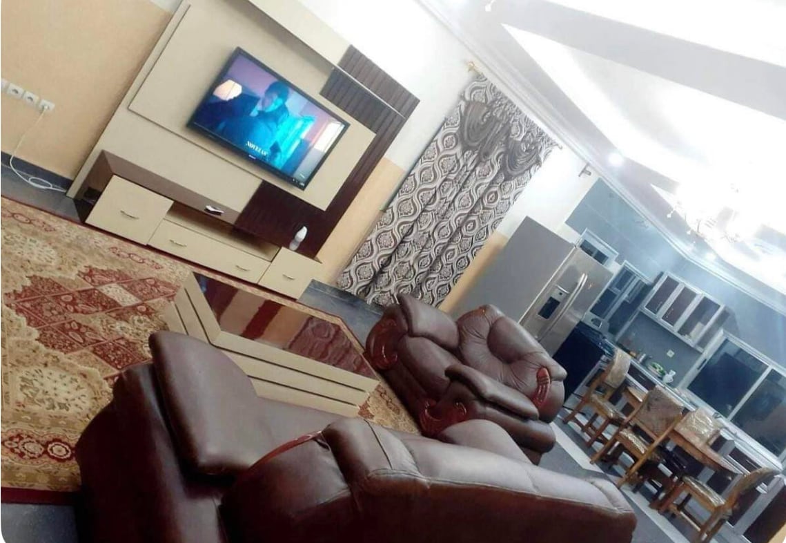 A spacious living area is illuminated by soft ceiling lights, featuring a large television mounted on a cream-colored media unit. Comfortable dark brown leather sofas surround a coffee table situated on a patterned area rug. An adjacent dining space is visible, with wooden chairs and a glass table.