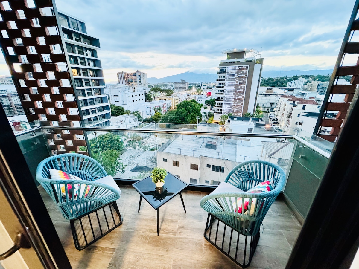 A balcony with a view features two modern blue chairs and a small black table. A potted plant is centered on the table, while the city skyline and distant mountains can be seen through large glass panels.
