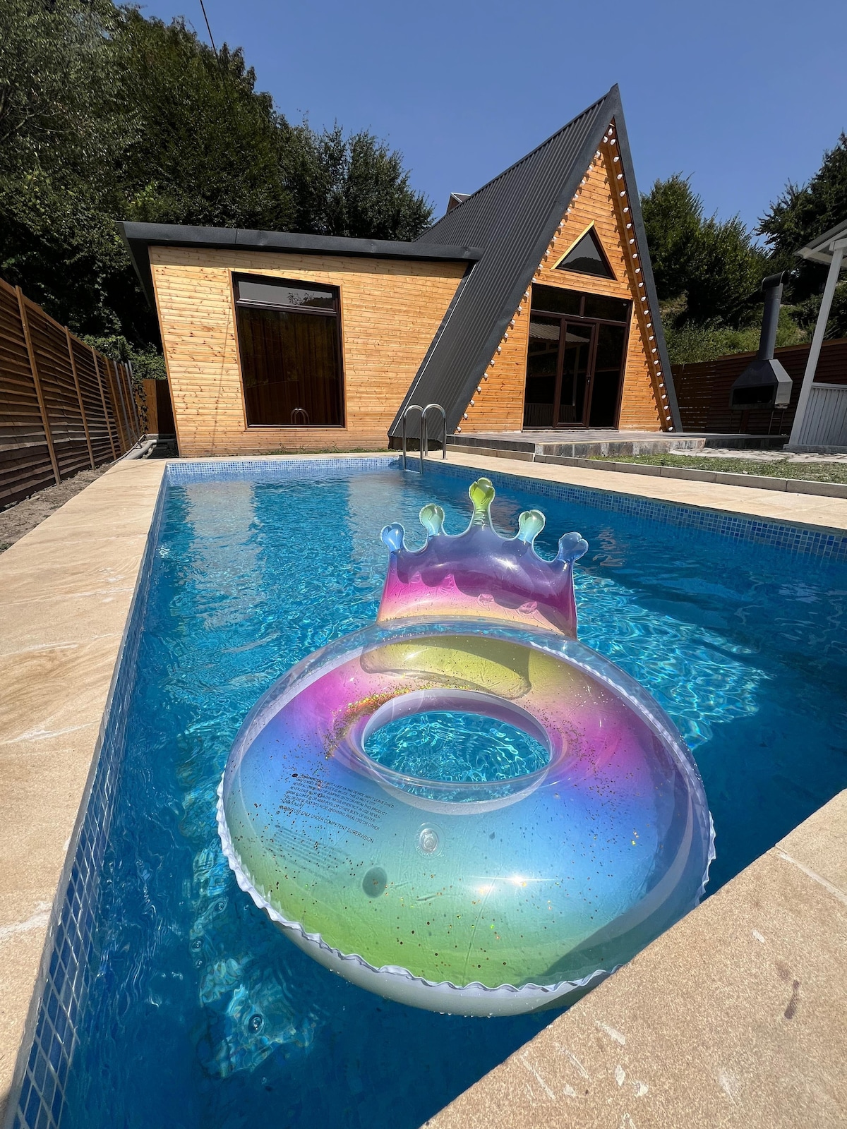 A modern cabin with a unique A-frame design is situated beside a clear swimming pool. A colorful inflatable pool float with a crown design rests in the water, reflecting sunlight, while the surrounding wooden fence provides privacy.