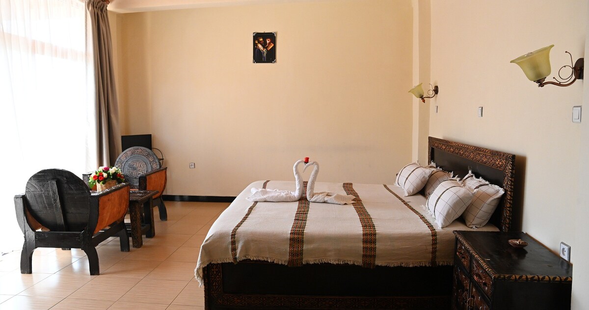 A spacious bedroom features a large bed adorned with a simple blanket. A pair of wooden chairs and a table is situated nearby, complemented by natural light filtering through a large window. Decorative lamps are mounted on the wall, enhancing the room’s layout.