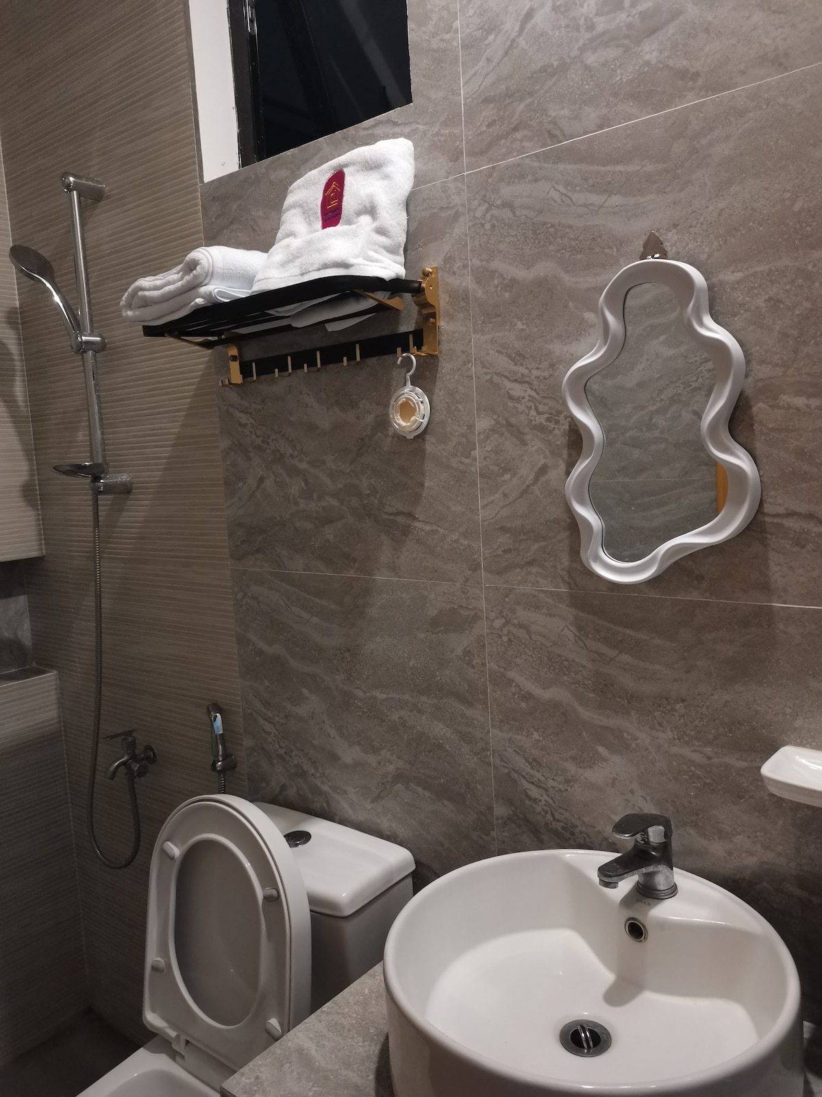 A modern bathroom features sleek gray tiles and a round sink atop a compact counter. Fresh towels are neatly arranged on a wall-mounted shelf, while a unique mirror adds a decorative touch. A shower area is equipped with a stylish faucet and a handheld showerhead.