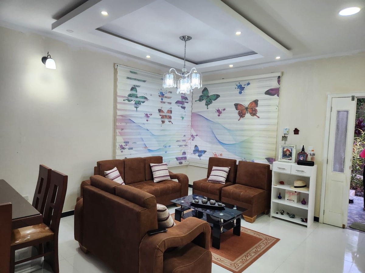 A welcoming living area features a set of brown upholstered sofas arranged around a central coffee table. Colorful butterfly patterns decorate the window shades, while a chandelier hangs from the ceiling. A dining table is visible in the background, enhancing the communal space.