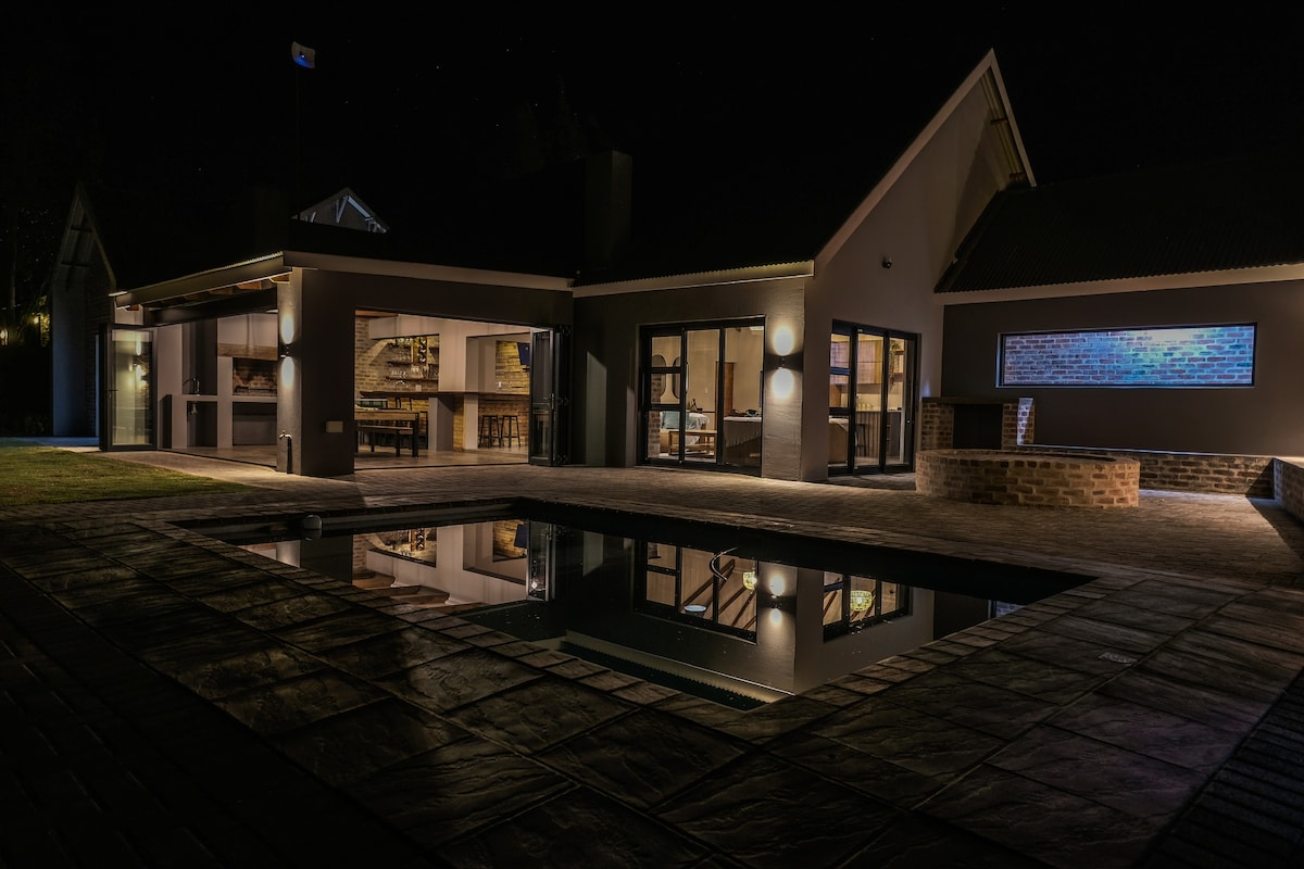 The exterior of the property is illuminated at night, showcasing large glass doors that open to a spacious patio and a pool. The pool's surface reflects the surrounding architecture and lights, highlighting the modern design elements and tranquil ambiance.