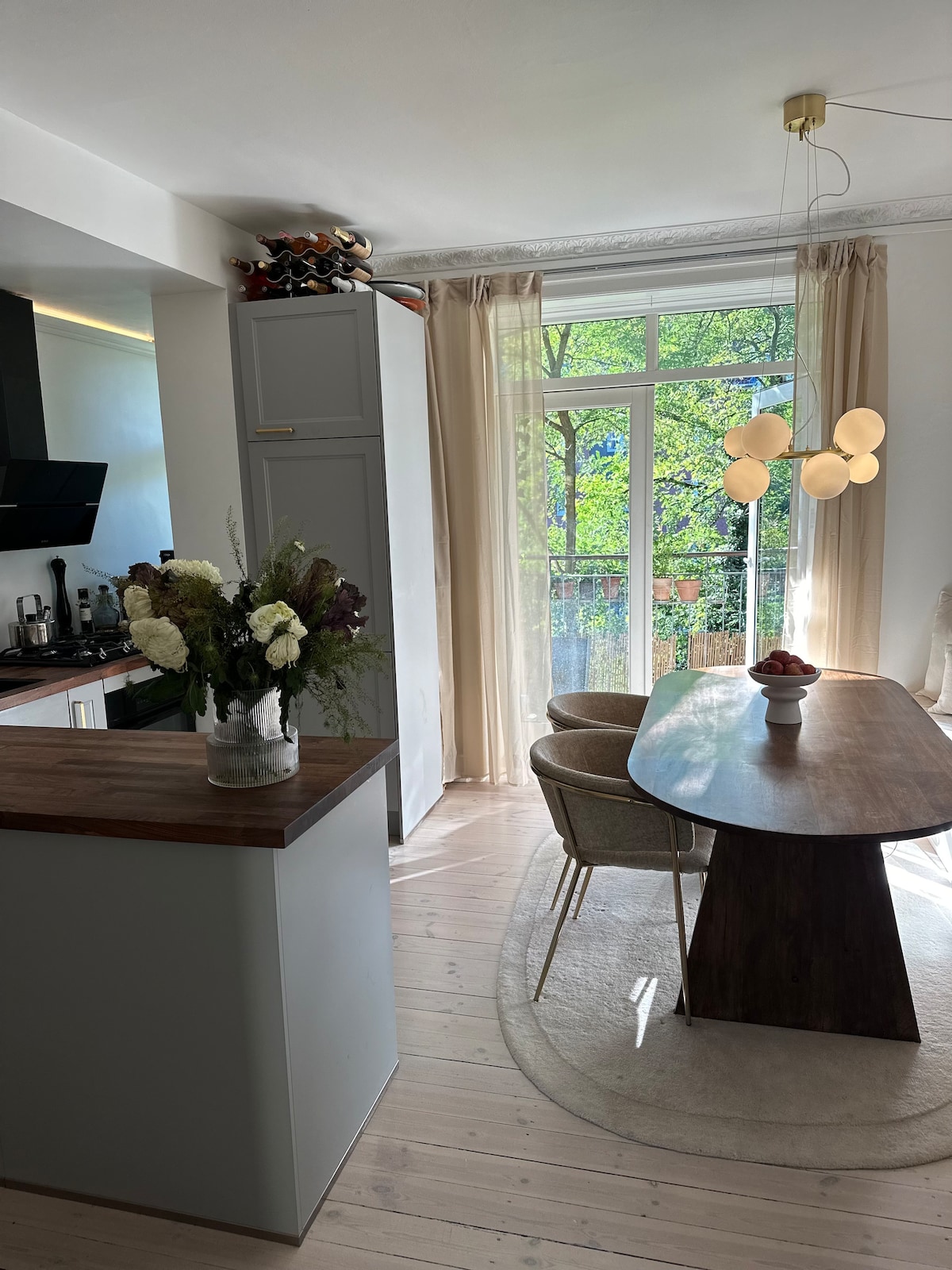The modern kitchen features sleek cabinetry and a spacious dining area. A large window provides natural light, showcasing leafy greenery outside. A wooden dining table is complemented by two comfortable chairs, while a vase of flowers adds a fresh touch to the setting.
