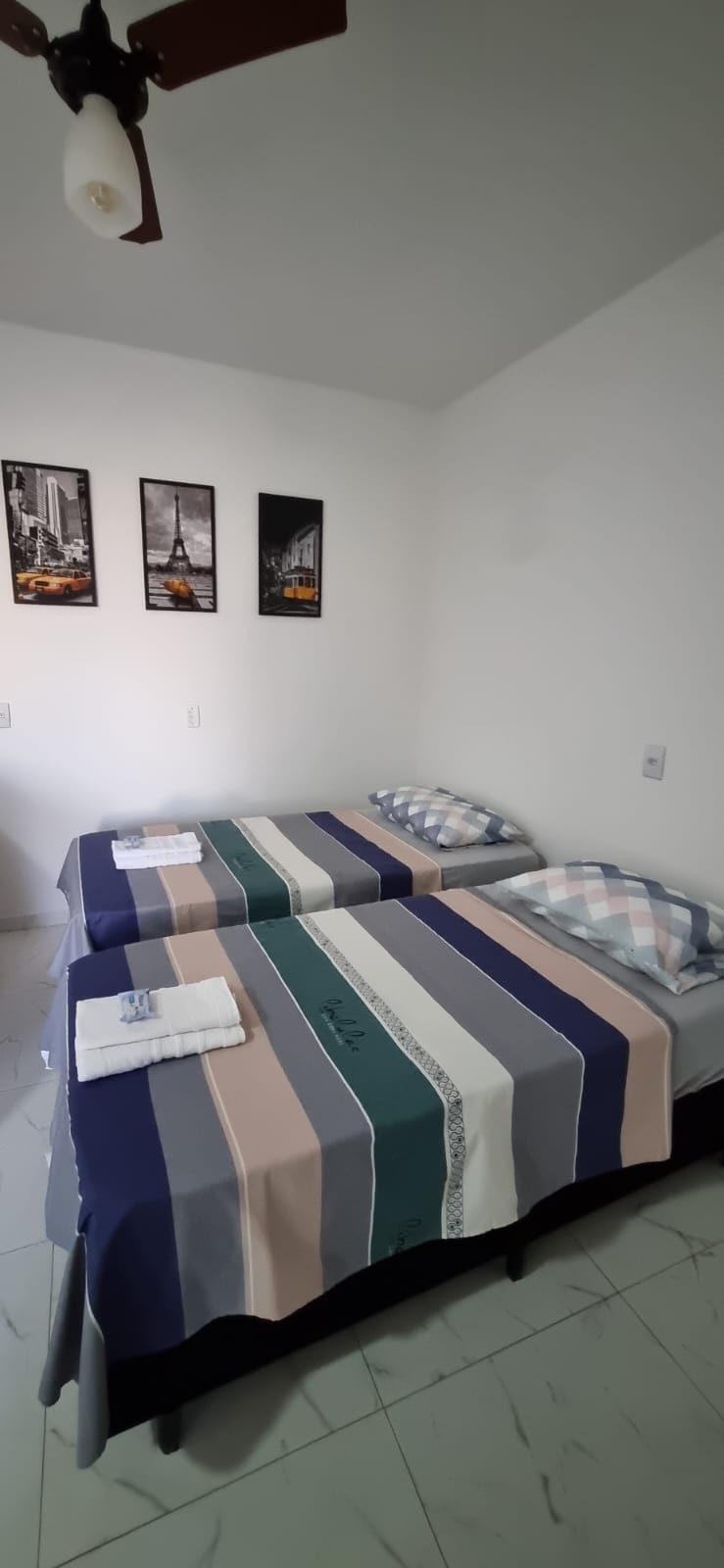A well-lit sleeping area features two neatly made beds, each dressed in coordinating striped bedding. Wall-mounted artwork depicting cityscapes adds a touch of character. Fresh towels are placed at the foot of each bed, contributing to a tidy and welcoming ambiance.