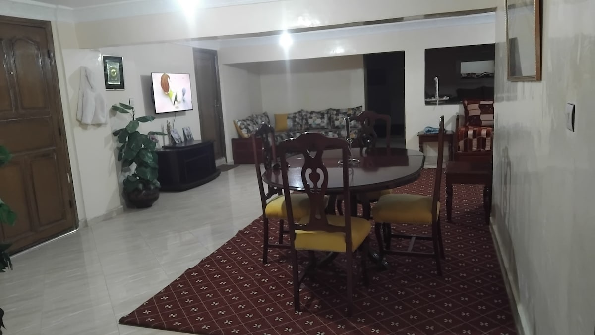 A spacious living area is presented with a dining table surrounded by yellow chairs. A comfortable seating arrangement is visible in the background next to a television. Natural light enhances the room's open layout, with decorative plants adding a touch of greenery.