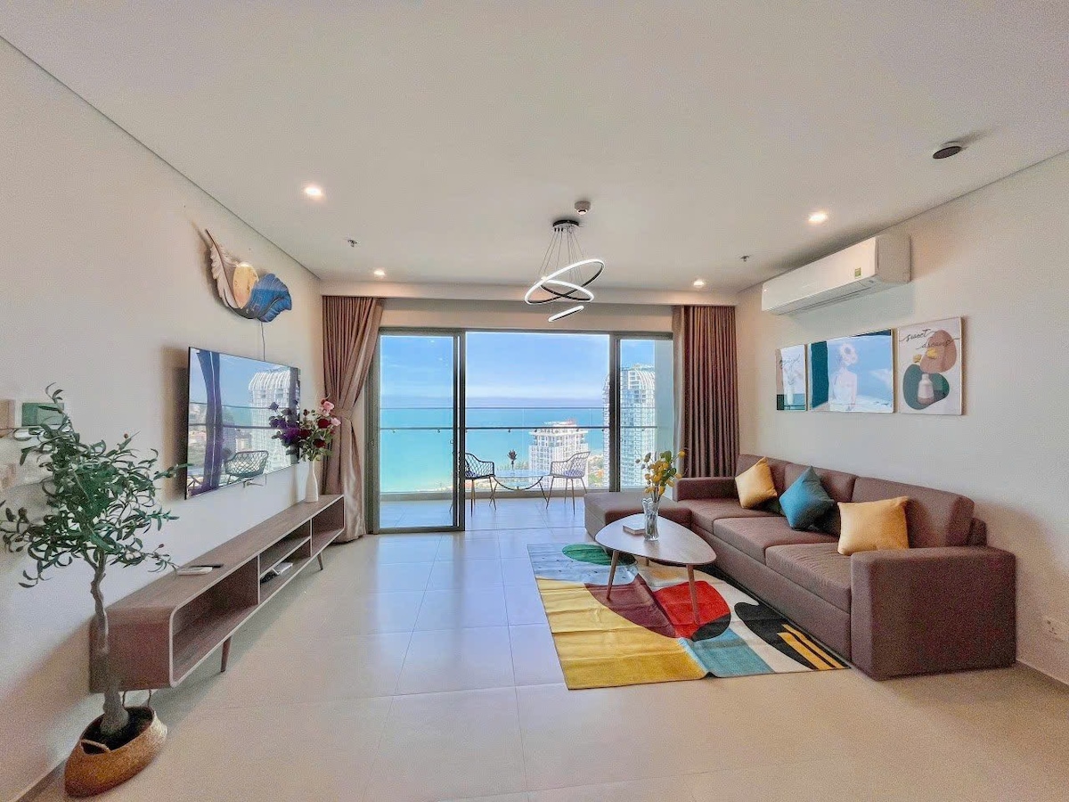 A spacious living room is presented with a large sofa, complemented by decorative cushions. A modern coffee table is centered on a colorful area rug. Glass doors open to a balcony with ocean views, allowing natural light to fill the room.