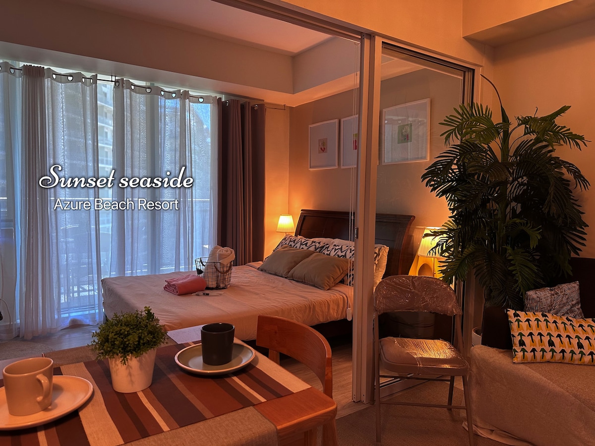 An inviting bedroom features a spacious bed adorned with patterned pillows. A curtain-draped window allows soft natural light to fill the room. A small table with plates and cups is positioned in the foreground, complemented by a potted plant adding a touch of greenery.