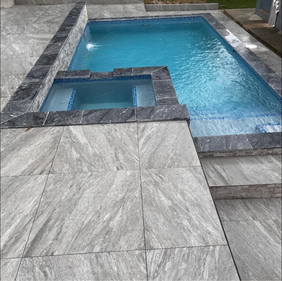 A clean, modern pool area is presented with tranquil blue water surrounded by grey marble tiles. The pool features a built-in spa with clear water and steps leading in, offering a seamless transition from the surrounding outdoor space.
