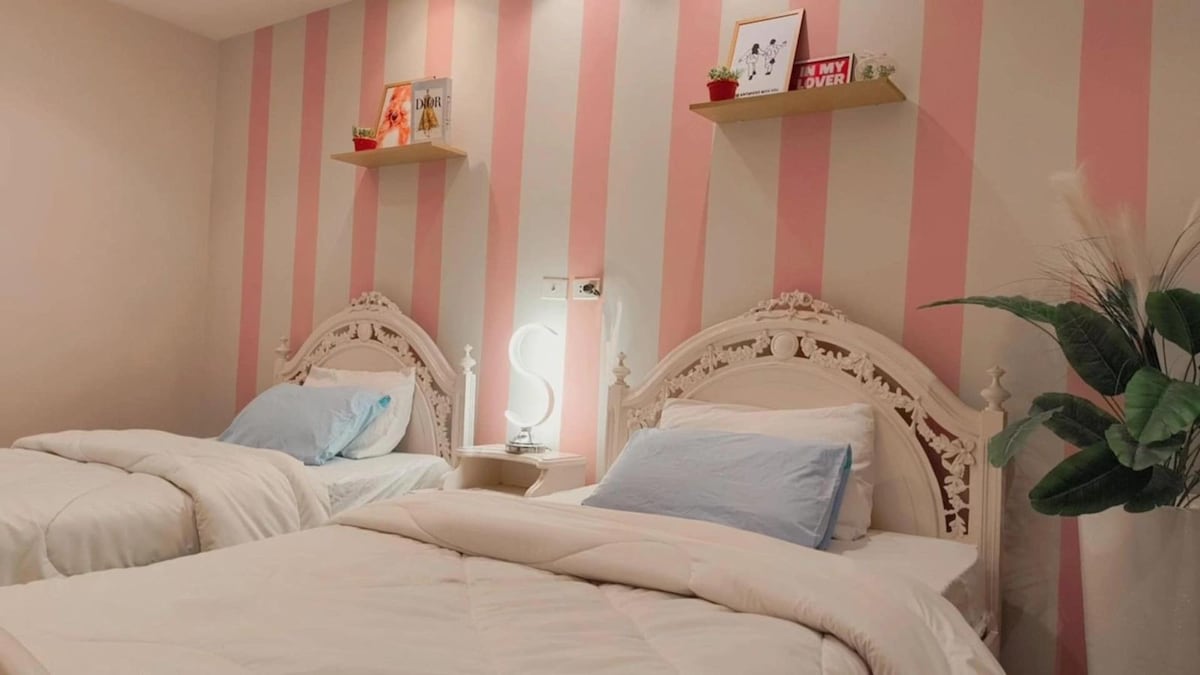 A bright bedroom features two spacious beds with light blue pillows and white bedding. Decorative shelves above hold small plants and framed items. A bedside lamp provides soft light, while a leafy green plant adds a touch of nature to the inviting space.