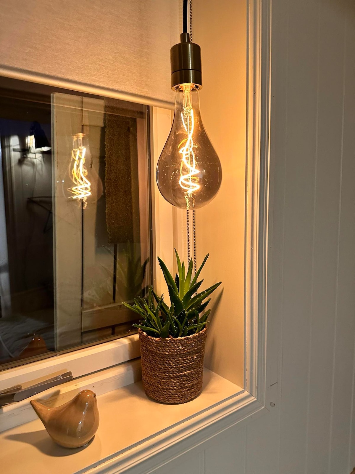A decorative corner features a vintage-style hanging light emitting a warm glow. Below, a potted succulent is placed in a textured basket, alongside a small ceramic bird. Reflections can be seen in the window, enhancing the overall ambiance of the space.