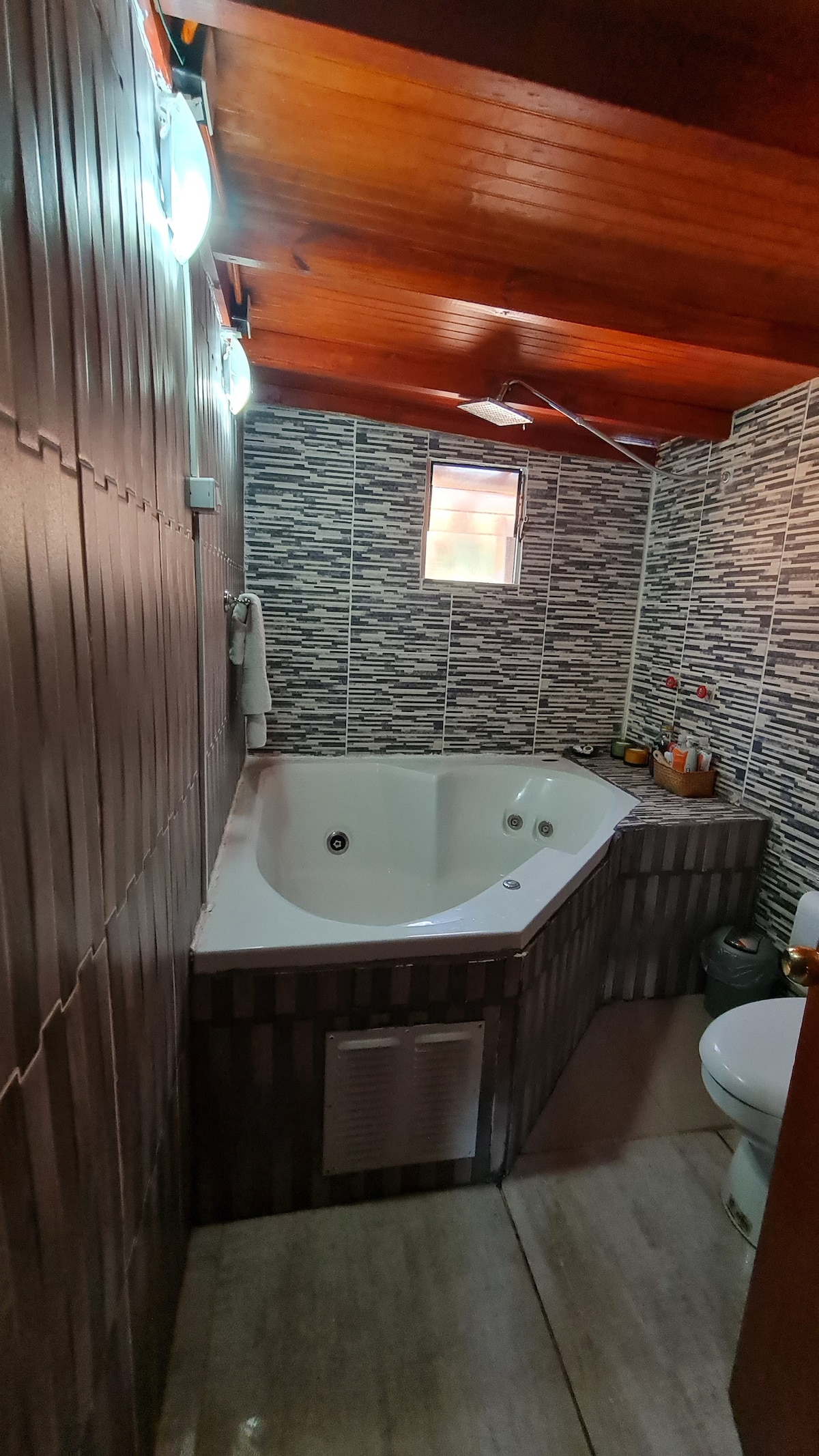 A spacious bathroom features a corner jacuzzi, offering a relaxing retreat. Textured wall tiles create an interesting backdrop, complemented by wooden ceiling beams. Soft lighting is provided by modern fixtures, and a small window allows natural light to enter, enhancing the room’s overall ambiance.