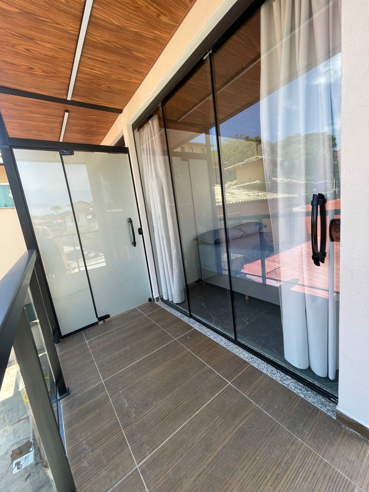 A private balcony is presented, featuring sliding glass doors and a wooden ceiling. The tiled flooring complements the space. Sheer curtains are drawn to the side, allowing for natural light, while a glimpse of outdoor scenery is visible through the transparent doors.