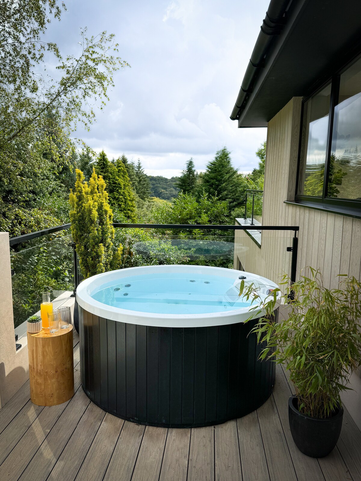 A private hot tub is positioned on a wooden deck, offering scenic views of the surrounding trees. The tub is circular, with a light blue interior and surrounded by a clear glass railing. A small wooden table with a drink rests nearby, adding a touch of comfort.