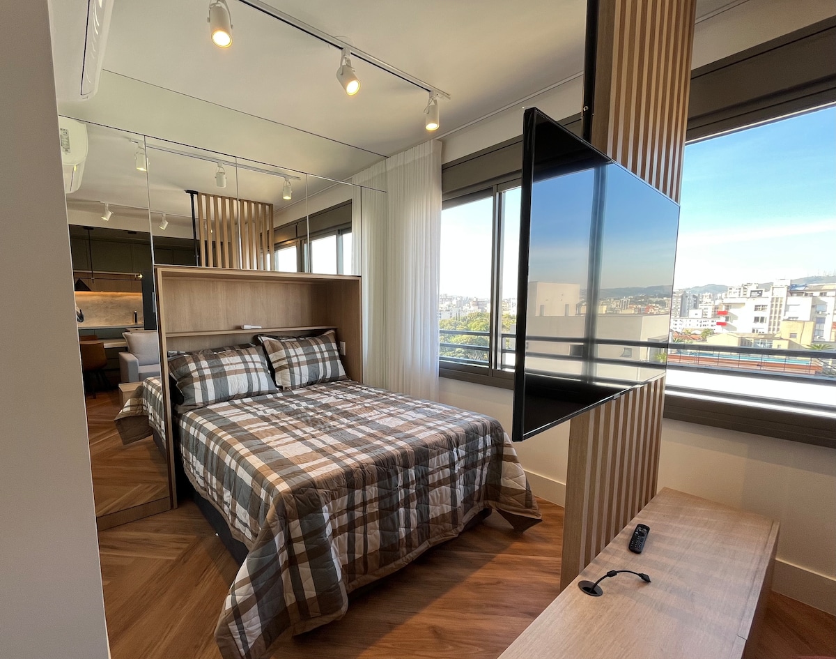 A cozy sleeping area features a checkered bedspread on a spacious bed. The room is illuminated by natural light from large windows, with a mounted television across from the bed. Wooden accents add a touch of warmth to the design.