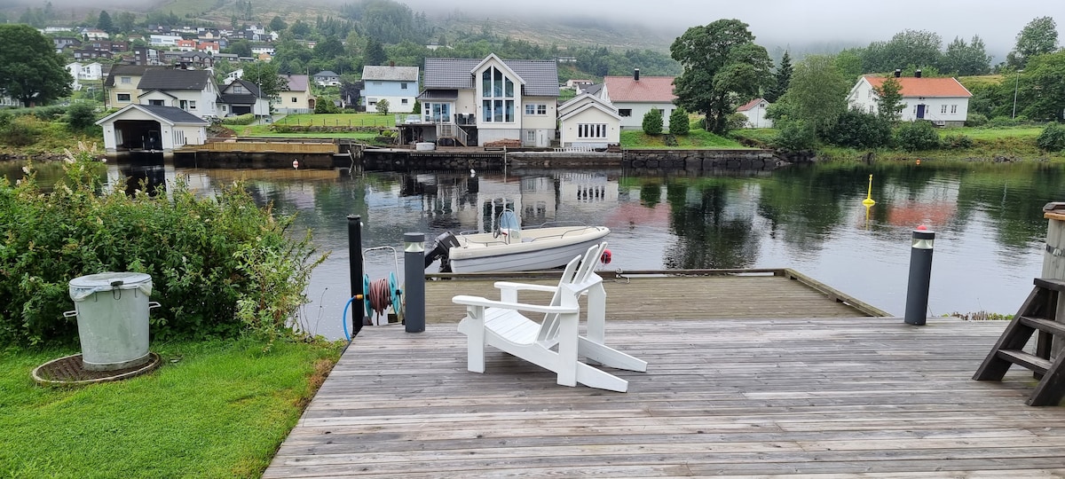 A spacious outdoor deck features white adirondack chairs overlooking a calm waterway. A small boat is moored nearby, while greenery frames the scene. Houses can be seen across the water, set against a backdrop of gentle hills and low-hanging clouds.